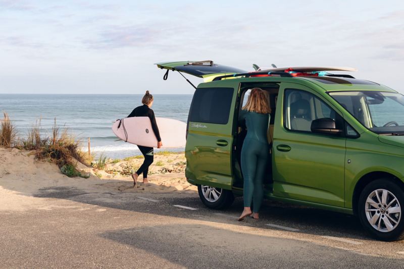 VW California vert devant la mer avec 2 femmes qui font du surf