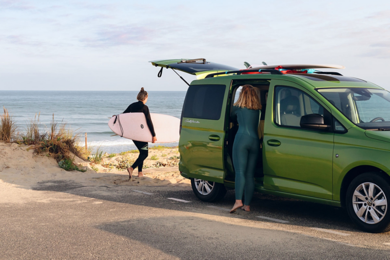 VW California vert devant la mer avec 2 femmes qui font du surf