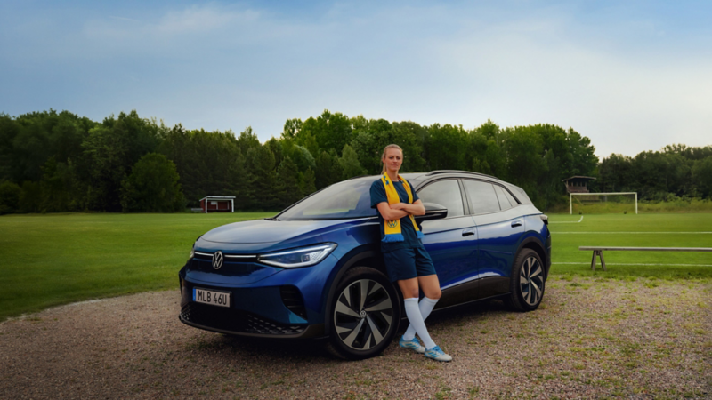 En blå VW ID.4 parkerad vid en fotbollsplan, sedd snett framifrån. Fotbollsspslare Amanda Ilestedt står lutad mot bilen