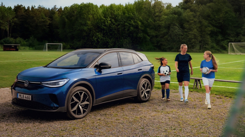 En blå ID.4 parkerad vid en fotbollsplan, en fotbollsklädd kvinna och två barn går tillsammans förbi bilen med en fotboll i handen