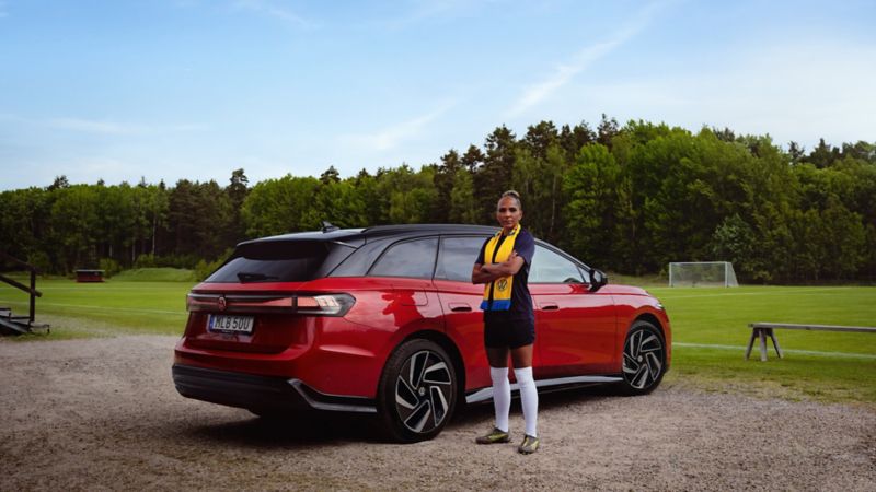 En röd VW ID.7 Tourer parkerad vid en fotbollsplan, sedd snett bakifrån. Fotbollspelaren Madelen Janogy står framför bilen