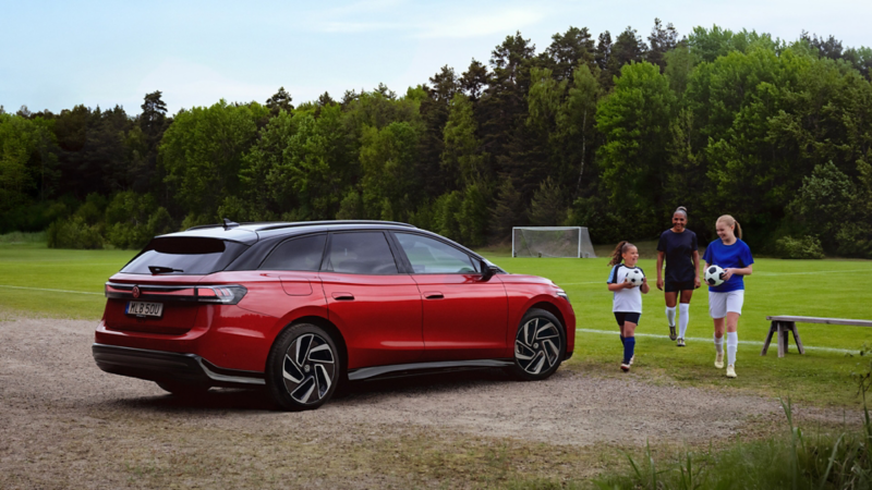 En röd ID.7 Tourer parkerad vi en fotbollsplan. En fotbollsklädd kvinna och två barn går förbi bilen med en boll