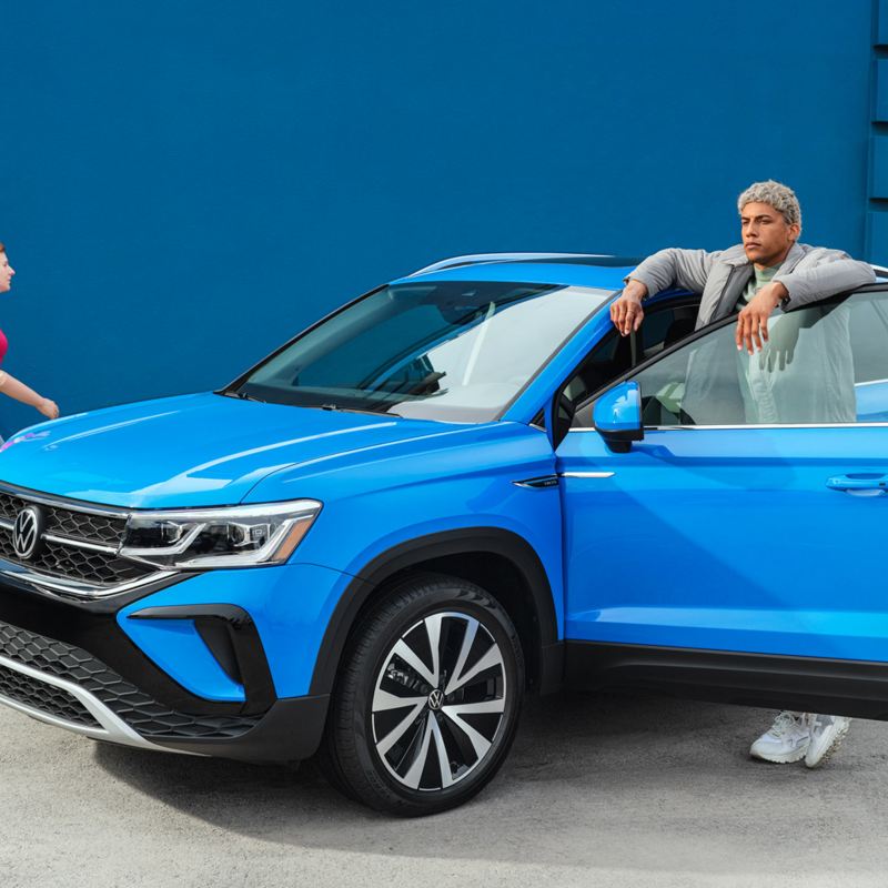 A man standing behind an open door of the Blue Metallic Volkswagen Taos while a woman is walking next to the car., link out to VW’s “new vehicle” category page