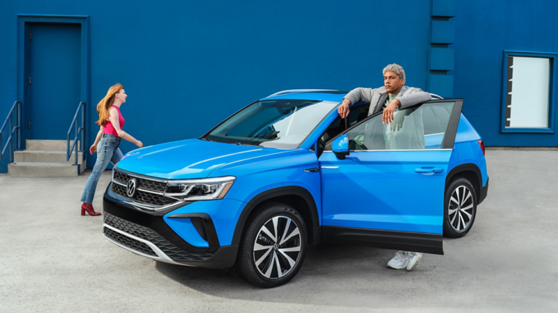 Un homme se tient derrière la portière ouverte d’un Taos de Volkswagen bleu métallisé pendant qu’une femme marche à côté du véhicule., lien vers la page vehicules-neufs
