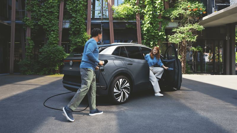 A man plugs in a pure gray electric VW ID.4, while a woman exits the car parked by a building adorned with green leaves