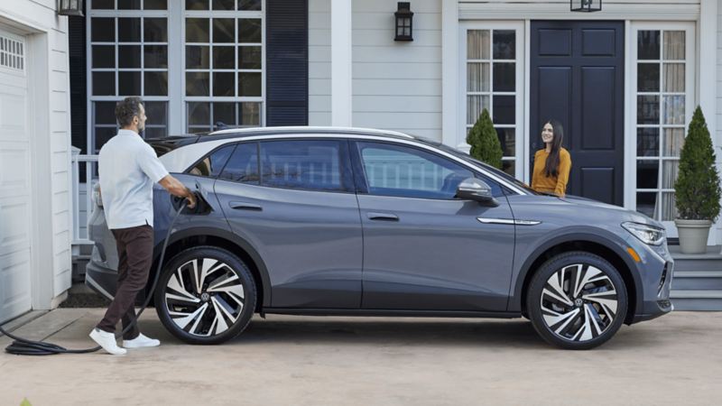 Un homme branche un ID.4 de VW gris pur à une borne de recharge à domicile, tandis qu’une femme se tient à côté du véhicule qui est stationné près d’une maison blanche.
