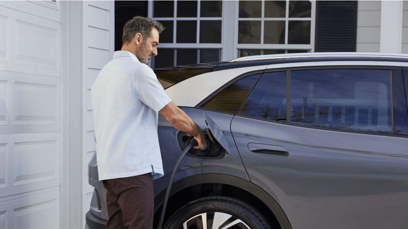 A close-up of a man plugging in a pure gray VW ID.4 parked by a white house