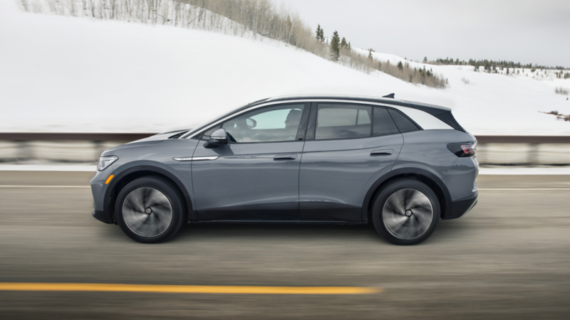 A gray Volkswagen car driving on a snowy road. Rolling hills and snow-covered trees are visible in the background