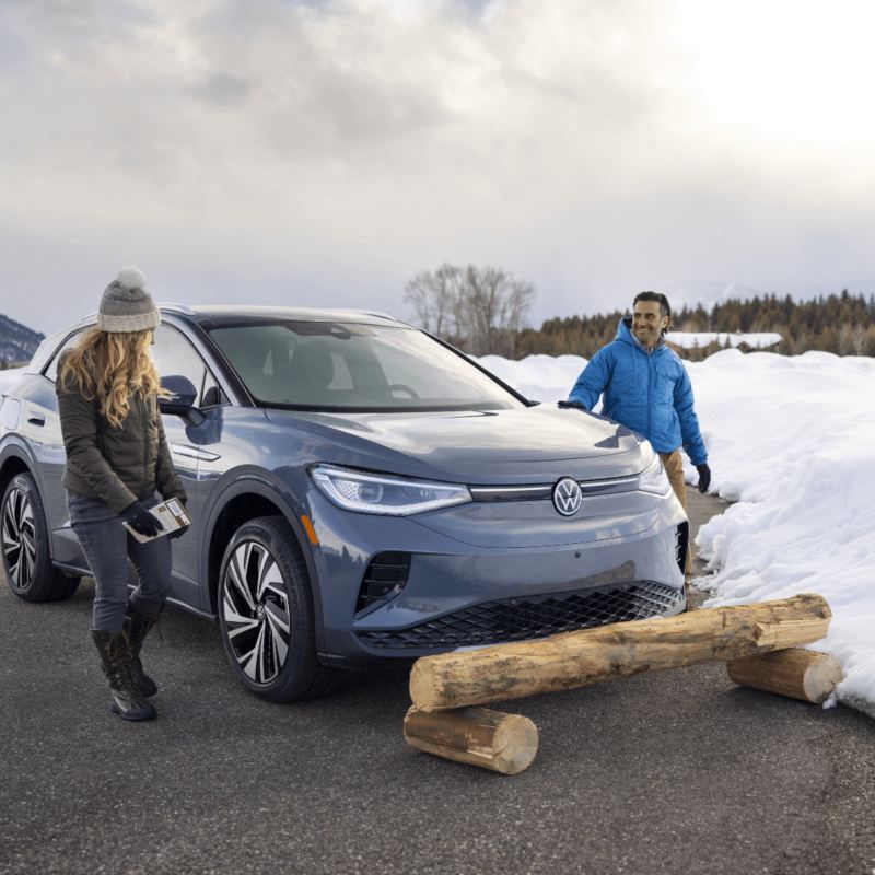 A grey Volkswagen SUV is stationed near a wooden log on a snow-covered road, while two individuals stand nearby against a backdrop of rolling hills and lush trees