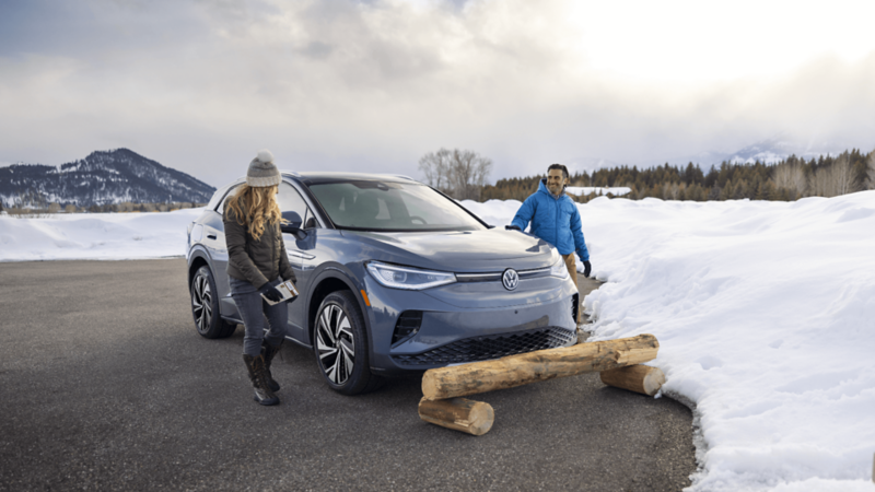 A grey Volkswagen SUV is stationed near a wooden log on a snow-covered road, while two individuals stand nearby against a backdrop of rolling hills and lush trees