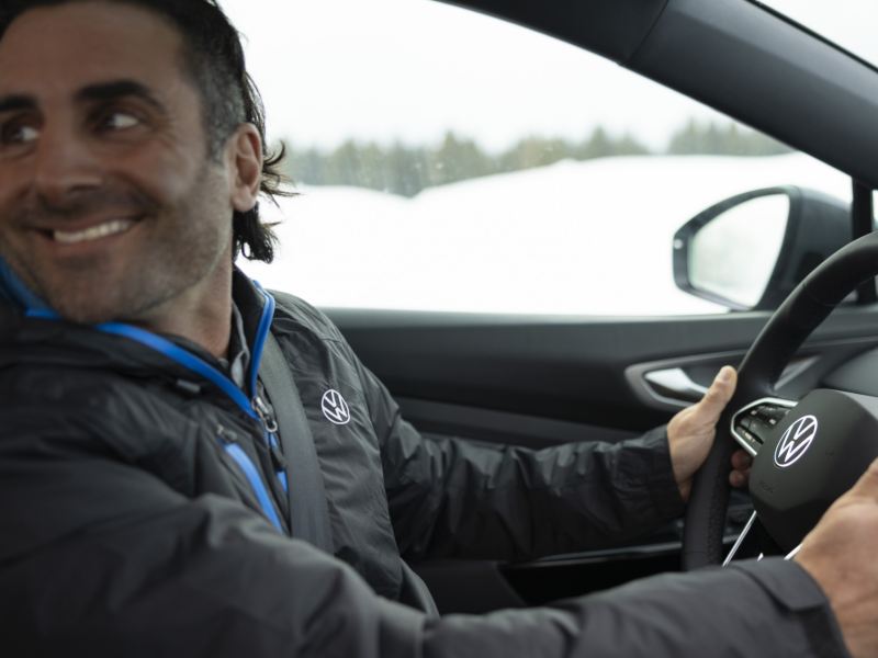 A man sits behind the steering wheel of a Volkswagen, glancing backward, with snowy hills adorned with pine trees in the background