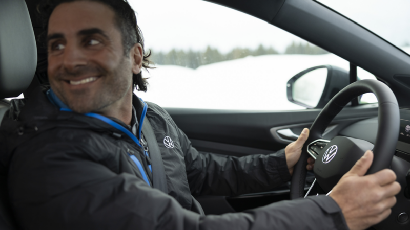 A man sits behind the steering wheel of a Volkswagen, glancing backward, with snowy hills adorned with pine trees in the background