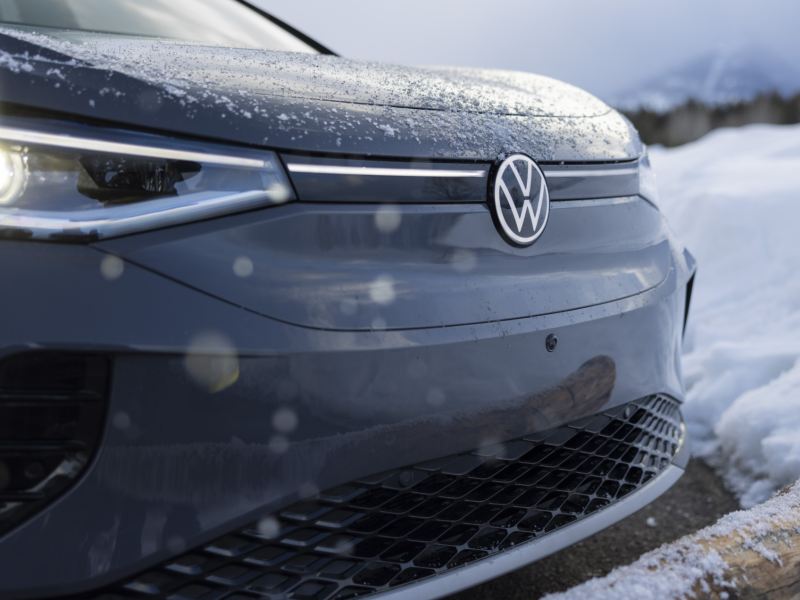 A close-up of the front end of the Volkswagen covered in light snow, parked next to a snowy terrain with mountains in the background
