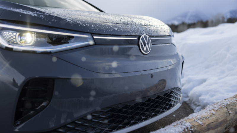 A close-up of the front end of the Volkswagen covered in light snow, parked next to a snowy terrain with mountains in the background