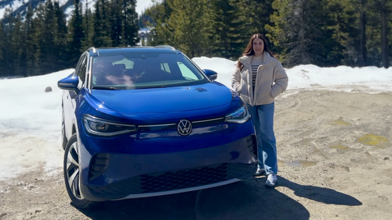 Une femme se tient près d’un VUS de Volkswagen bleu stationné sur une route de gravier avec de la neige et des pins en arrière-plan.