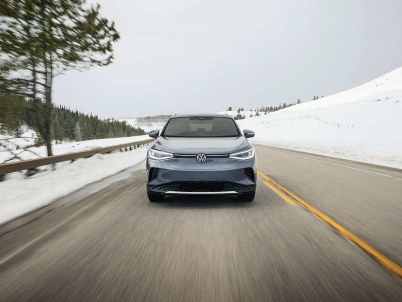 Vue frontale d’un VUS de Volkswagen gris qui roule sur une route de montagne enneigée, flanquée de collines enneigées et de pins.