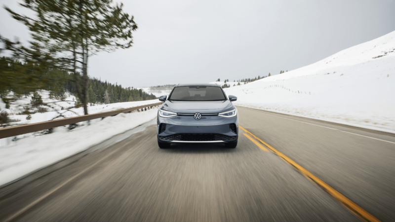 A frontal view of a gray Volkswagen SUV driving along a snowy mountain road, flanked by snow-covered hills and pine trees