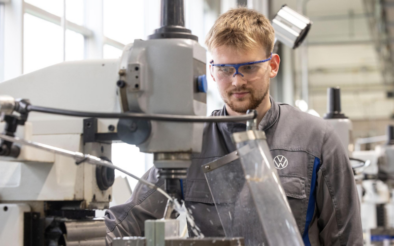 VW Auszubildender in Schutzkleidung arbeitet in einer Halle an einer Maschine