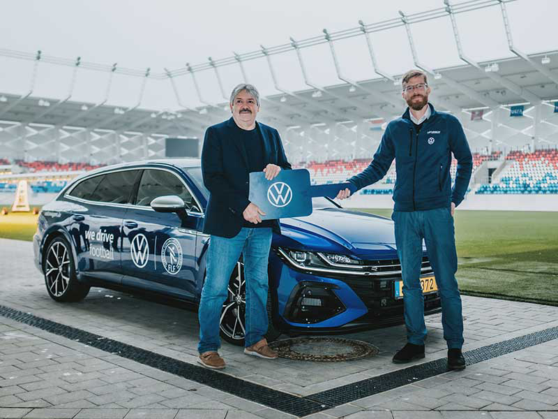 Deux hommes posent souriants devant une Volkswagen bleue dans un stade, l'un d'eux tenant une grande clé ornée du logo VW. Cette scène symbolise le partenariat entre Volkswagen Luxembourg et la Fédération Luxembourgeoise de Football (FLF).