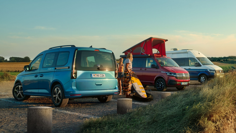 vw Caddy California, vw california 6.1 avec toit relevé et Grand California garés sur un parking dans un paysage près de la mer avec des surfeurs et promeneurs