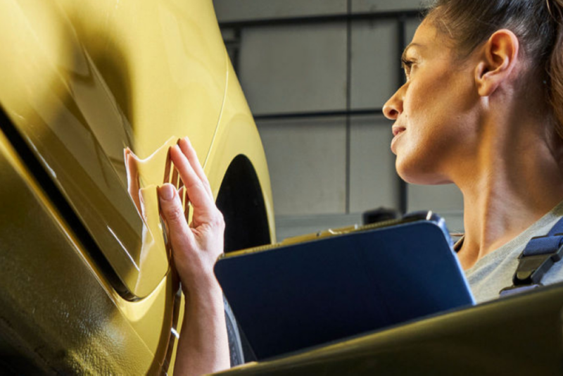 A woman in blue overalls inspects a golden VW vehicle, holding a clipboard.