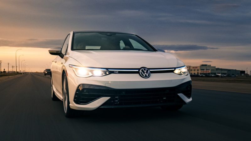 Une Volkswagen Golf R 2024 blanche roule sur une route au crépuscule, mettant en valeur son module d'éclairage d'ambiance. Le ciel en arrière-plan présente un dégradé de couleurs allant du bleu à l'orange.