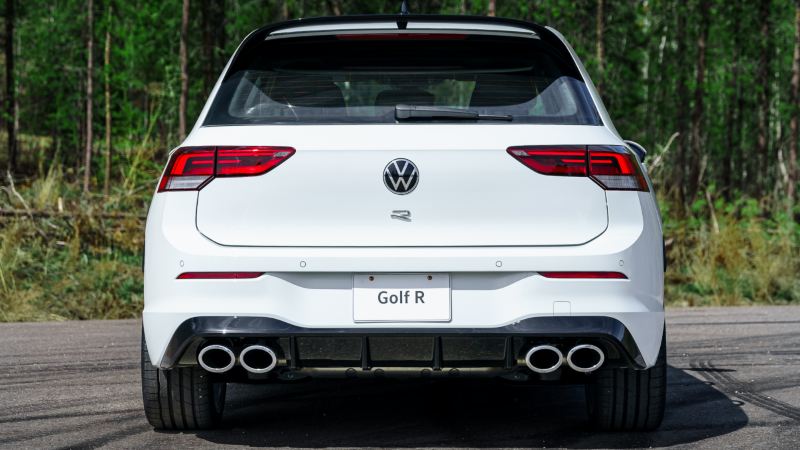 A rear view of a white 2024 Volkswagen Golf R parked on a road with lush greenery in the background.