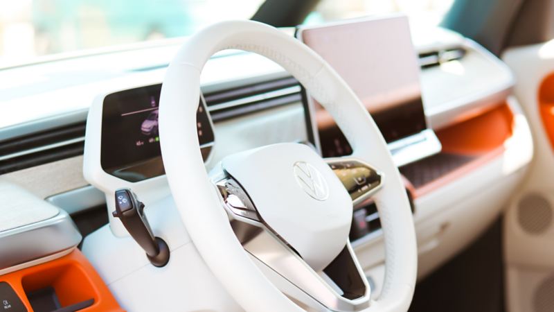 L'intérieur d'un Volkswagen ID.Buzz présente un volant blanc avec un emblème argenté au centre, un tableau de bord orange et blanc, et un écran d'affichage numérique derrière le volant.