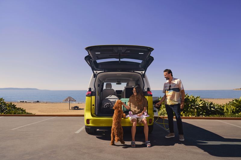 Vater, Tochter und Hund stehen am Strandboulevard vor der geöffneten Heckklappe des neuen VW ID. Buzz.