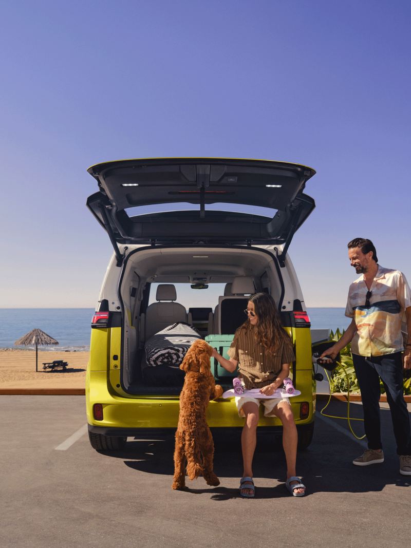 Ojciec, córka i pies stoją na bulwarze przy plaży przed otwartą tylną klapą nowego VW ID. Buzz.