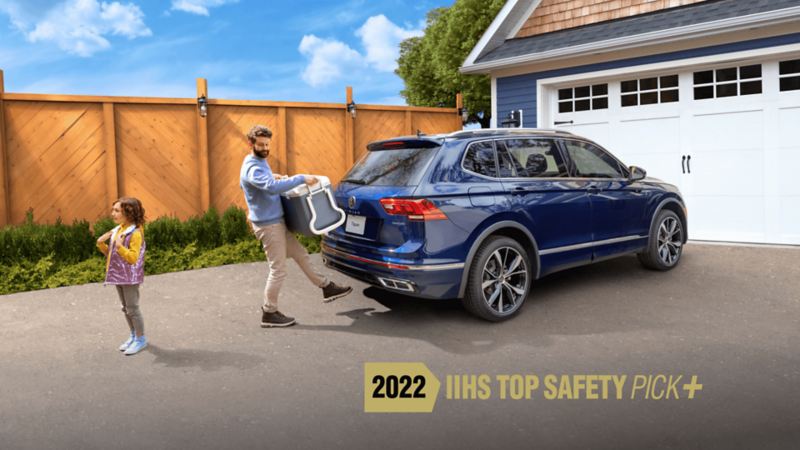 A man holding large cooler standing beside his daughter puts his foot under the back of 2022 Tiguan to activate the Easy Open  tailgate. 