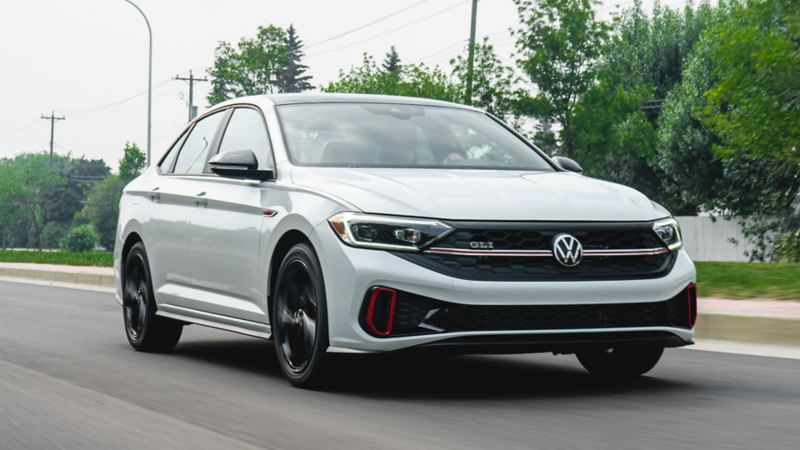A white 2023 Volkswagen Jetta GLI driving on a road with greenery in the background.