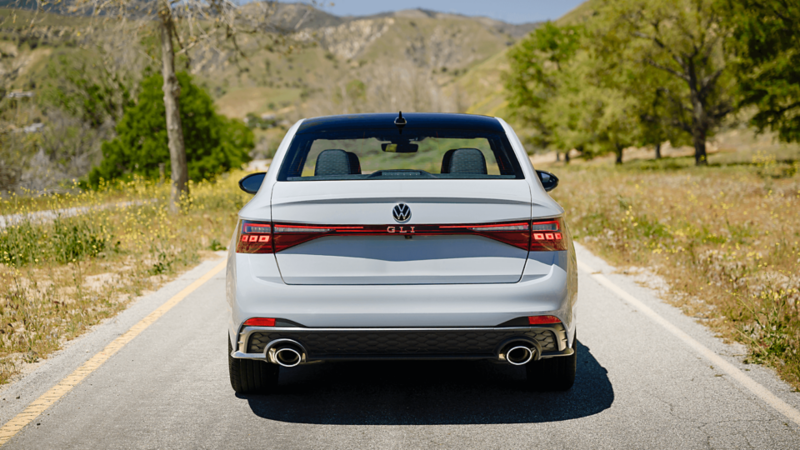 Rear view of a white 2025 Jetta GLI parked on a scenic road with grassy hills in the background.