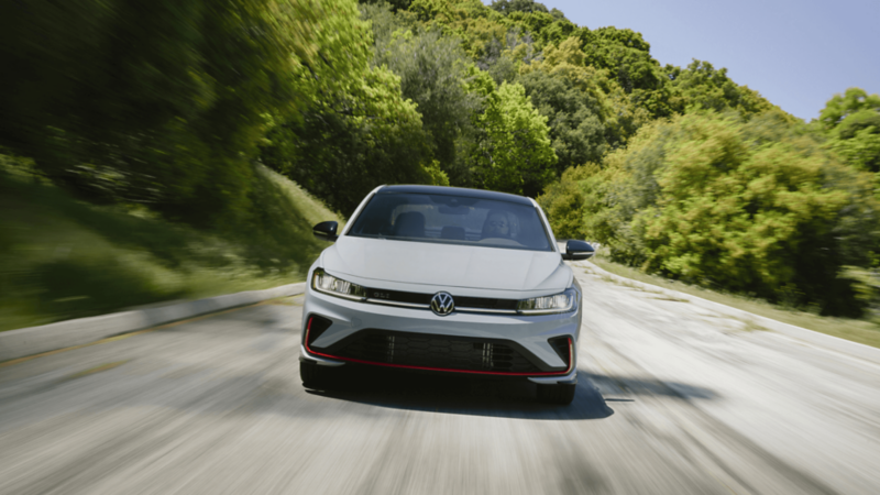 A silver Volkswagen Jetta GLI 2025 in motion on a road, with blurry mountain hills in the background.