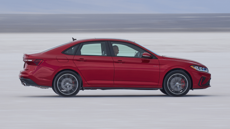 Side view of the 2026 Volkswagen Jetta GLI in red, driving across a white salt flat with a clear sky backdrop.