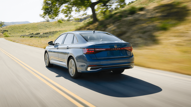 Rear view of the blue Jetta 2025 driving on a scenic road with a large tree and grassy hills in the background. 