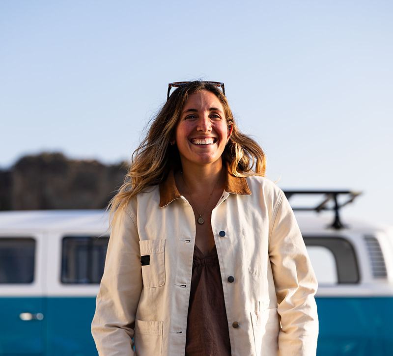Woman smiling in front of a retro blue Volkswagen bus with a cliffside and blue sky in the background