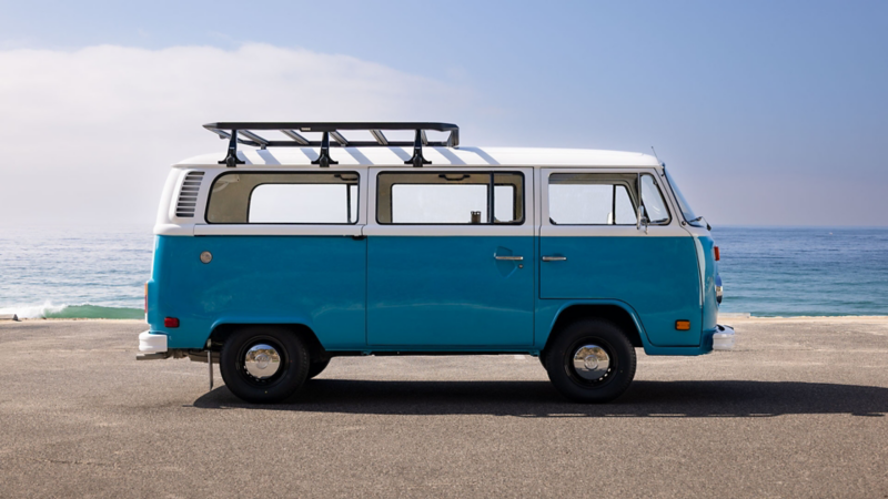 A retro blue Volkswagen bus with a roof rack parked in an oceanside lot on a sunny day