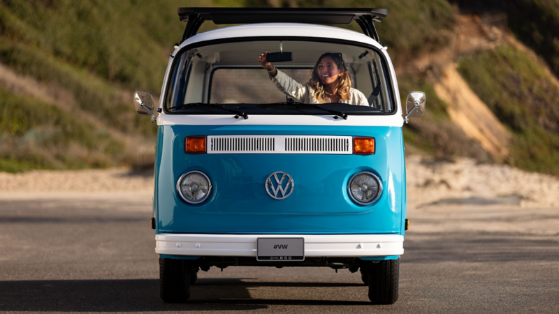 Head-on view of a woman adjusting the interior rearveiw mirror from the front seat of a retro blue Volkswagen bus parked in a cliffside lot