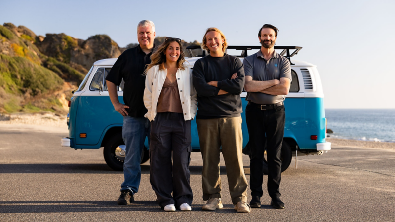 Four smiling people standing in front of a retro blue Volkswagen bus parked in a cliffside lot by the beach