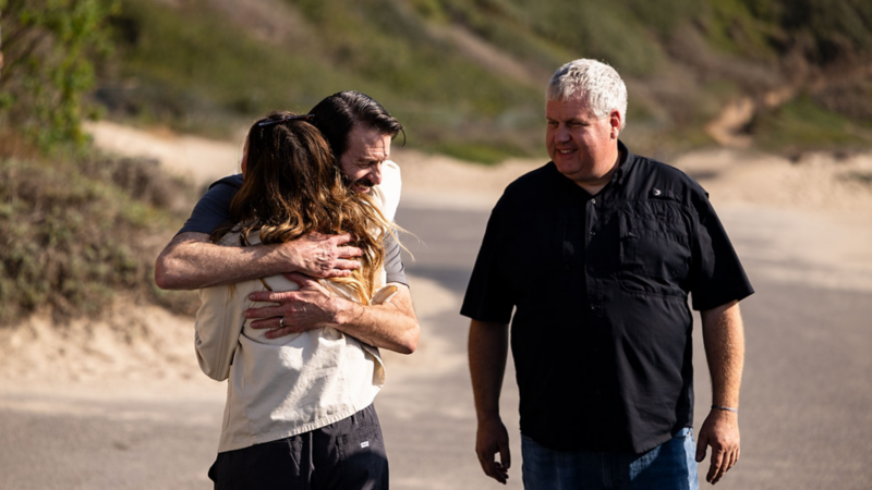 Woman graciously hugging a man near another man smiling with cliffsides in the background