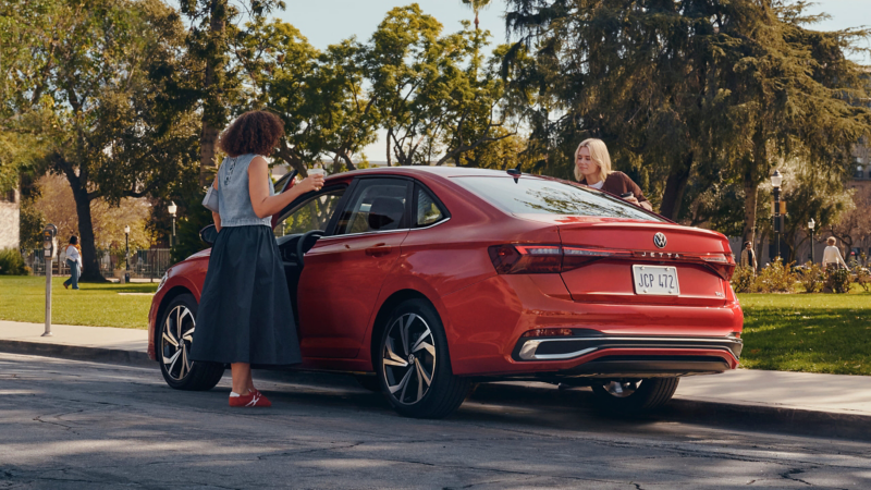 Dos mujeres con tazas de café se suben a un Jetta que se muestra en Kings Red Metallic parqueado en un parque exterior bordeado de árboles.