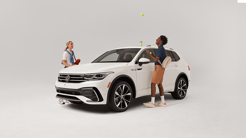 Vista frontal de 3/4 de un Tiguan que se muestra en blanco ópalo estacionado sobre un fondo blanco con dos personas paradas afuera del vehículo jugando con equipo de tenis.