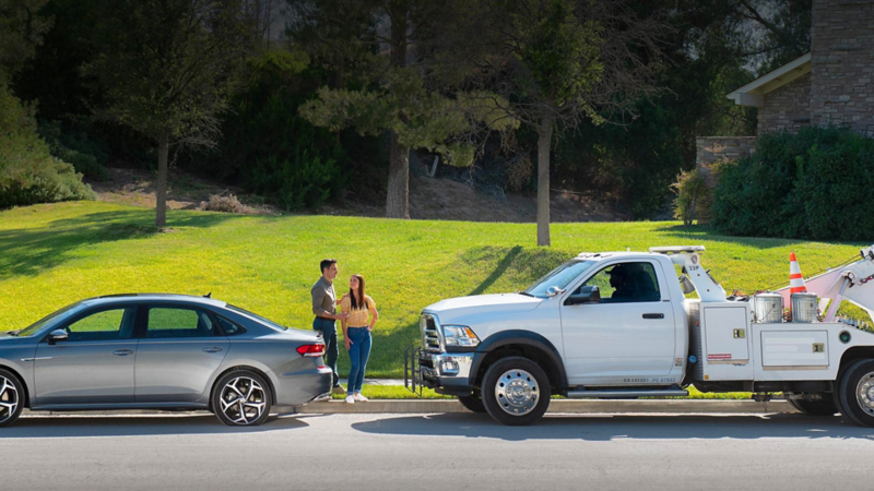 A Volkswagen with a rear flat tire is parked on the side of a road. A couple stands near the trunk facing an approaching VW Roadside Assistance service truck.