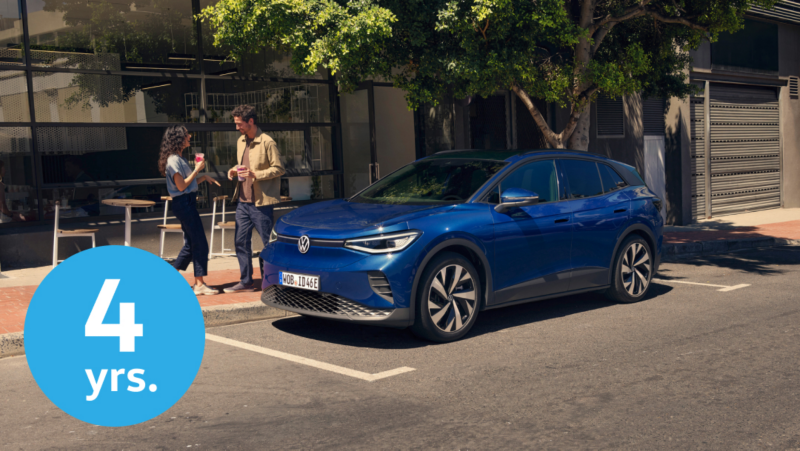 Un Volkswagen ID.4 azul está estacionado en una calle de la ciudad frente a un café donde dos personas conversan con tazas en la mano, y hay un gran círculo azul con el texto "4 años" en la esquina inferior izquierda.
