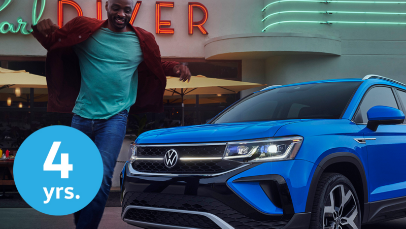 Close-up shot of a man walking in front of a Taos in Cornflower Blue parked in front of a neon-accented restaurant named “Pearl Diver.”