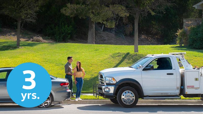 Un Volkswagen con una rueda trasera pinchada está aparcado en el arcén de una carretera. Una pareja se encuentra cerca del maletero frente a un camión de servicio de asistencia en carretera de VW que se aproxima. En la esquina inferior izquierda de la imagen hay un icono circular azul claro con un número y un texto que dice "3 años".