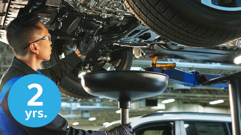 A VW service technician stands underneath a Volkswagen that is on a car lift, draining the old oil out and into a catch pan. In the lower left corner of the image is a light blue circular icon with a numeral and text reading “2 years.”
