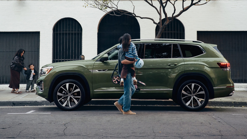 Un Atlas en color Avocado Green Metallic estacionado frente a un edificio verde, mientras un papá levanta a su hijo para que toque la puerta trasera abierta.