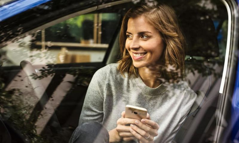 Smiling woman sits in the front seat of a parked VW, smartphone in hand.